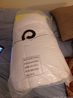 Rolled and packaged white mattress topper in plastic wrap placed on a bed with pillows.
