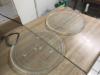 Two transparent round glass microwave trays placed on a wooden surface showing their flat shape and three central raised fitting points.