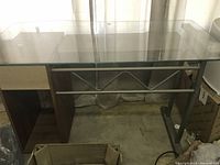 Front view of office desk showing clear glass tabletop, metal frame with geometric design, wooden side panels and slot drawer.