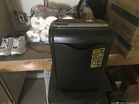 Front view of a black paper shredder sitting on a wooden platform in a storage area, showing the operational sticker and overall condition.