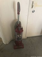 Full view of a red and gray Shark upright vacuum cleaner standing against a wall, showing the power cord and dustbin.