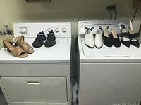 Six pairs of women's shoes lined on top of two white appliances (washer and dryer). Shoes include tan sandals, black shoes, and other assorted heels.