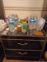 Wooden bedside table with two drawers. Various bottles and containers on top, not included in lot. Table positioned against wall with window blinds above.