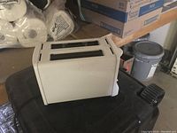 Photo of white two-slot toaster placed on a black surface in storage area with boxes visible in the background.