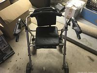 Photo showing a metal framed walker with padded black vinyl armrests, a foldable seat, and backrest in a storage area with boxes and a carpet roll nearby.