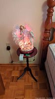 RGB light-up angel figurine displayed on small round wooden table against wall near bed frame and electrical outlet.
