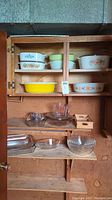 Overview of wooden shelves containing multiple Corning Ware dishes with floral and solid patterns on the upper shelves, and clear glass dishes and measuring cups on the lower shelves.