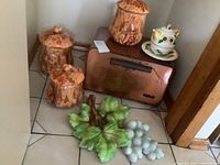 Photo showing bread box, three mushroom canisters, teapot and cup, and grape leaf wall hanging on tile floor.