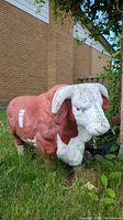 Left-side view of red and white cement bull statue placed on grass near a brick building showing weathered paint and overall condition