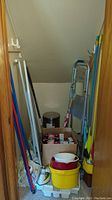 Photo of small closet space containing multiple colorful mop handles, yellow bucket with basins, metal folding step ladder, box with light bulbs, and small metal trash bin.