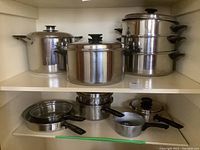 Photo showing multiple stainless steel pots with lids and several frying pans with black handles on shelf.