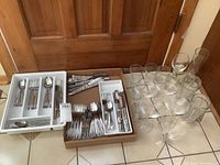Photo showing mixed sets of stainless steel cutlery in trays alongside various types of clear glassware including martini glasses, pilsner glass, milkshake glasses, tumblers, and a white wine glass.