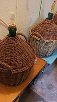 Full view of two large wicker wrapped demijohns with green glass necks and tubing inserted into cork stoppers, placed on wooden bench near a wall.