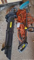 Photo showing a black blower vacuum and a black hedge trimmer with orange extension cords coiled beside them on a concrete floor.