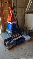 Full view of all items stacked in garage, including coolers at bottom, fishing rods standing vertically, tackle boxes, and portable barbecue grill.