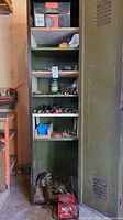 Green metal storage cabinet with shelves holding various hand tools and hardware parts including brushes, files, small containers, and other assortments.