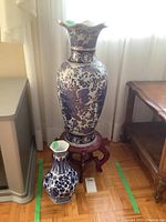 Pair of blue and white Asian style vases displayed on the floor next to furniture. Larger vase is on a wooden stand and smaller vase sits beside it.