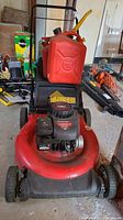 Front view of red Yard Machines gas powered push lawnmower with engine and two red gasoline cans placed on top.