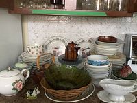 Overview of mixed dishware assortment including bowls, plates, green glass bowl in wicker basket, wooden pepper grinders, salt and pepper shakers, and teapots.
