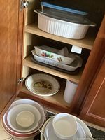 Inside kitchen cabinet showing multiple shelves with cookware including ceramic and glass casseroles, pie plate, and mixing bowls nestling inside each other