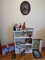 Full view of shelf with porcelain Nativity set, plates, statues, plaques and religious items alongside framed prints on wall and floor