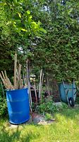 Full view of blue resin rain barrel filled with assorted scrap wood and wooden poles, green wheelbarrow and garden tools in the background.