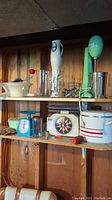 Wide image of wooden shelves holding vintage kitchen appliances and tools including two hand held mixers, milkshake maker, mechanical sifter, analog scales, sugar jar, and measuring cup.
