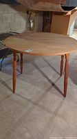 Round unfinished wood table with four turned legs and fold down sides shown in ambient lighting