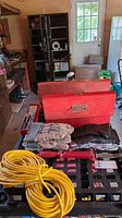 Two red metal tool boxes, multiple work gloves, an antique nail puller with red handle, socket set trays, and yellow extension cord on black crate surface.