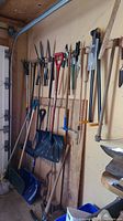 Wide view of various yard tools hanging on garage wall, including shovels, rakes, and clippers.