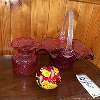 Photo shows three items: a ruffled cranberry glass basket with clear spiral handle, a hobnail cranberry glass vase, and a small painted ceramic candle bowl with lid decorated with yellow and white flowers on red.