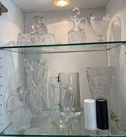 Two glass jugs, salt and pepper shakers, large vase, small vase, crystal decanter, and a clear glass tankard mug on bottom shelf inside glass cabinet