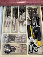 Cutlery drawer showing forks, knives, teaspoons with goose decor handles and various kitchen tools including scissors, peelers, corkscrews, and a bottle opener.
