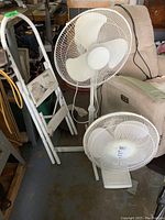 Photo showing large white Holmes electric fan on stand, small white table top fan, and white folding metal step stool.