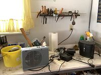 Wide view showing heater, bucket, hand tools hanging on rack, black battery adapter, and other miscellaneous items on table.
