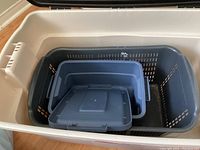 Large white Rubbermaid storage bin containing multiple plastic baskets including a dark gray basket with a lid and smaller blue basket inside it