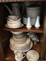 Photo showing coffee pot, stacked plates, bowls, cream pitcher, covered sugar bowl, and salt & pepper shakers in a wooden cabinet.