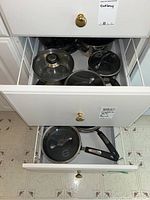 Open top drawer showing several pots with glass lids, black handles, stainless steel bodies.