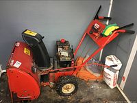 Full view of red MTD Yard Machines snow blower with 5 hp engine and 24-inch clearing width in a room. Includes gloves and bags nearby.
