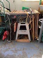 Full front view of TecoMaster table saw on metal stand, with folding wooden work bench on the right and reciprocating saw stored underneath.