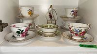 Full display shelf showing seven tea cups with matching saucers and one extra cup, all featuring floral patterns and gold trim.