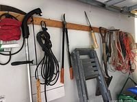 Overview of tools hanging on a wall including the electric weed eater, extension cords, pruners, shovel, machete-type blade, and garden hoe.