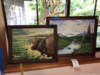 Two framed landscape paintings; one coastal cliff scene with waves, one forested mountain river scene
