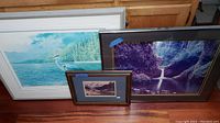 Three framed prints standing against a wooden cabinet: large heron print, medium waterfall print, smallest winter mountain scene.