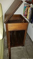 Side view of wooden music cabinet with hinged lid open, showing storage compartment and wooden partitions inside for records.