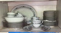 Shelved Noritake china including stacked plates, cups, saucers, an oval vegetable bowl, and an oval serving platter with blue floral and silver trim pattern.