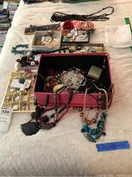 Photo showing assorted costume jewelry including various necklaces, pins, earrings, and accessories arranged on a bed with a pink box and organizational trays.