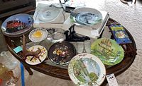 Top-down view of decorative plates arranged on oval glass-topped wooden table showing variety of designs including train, floral, and landscape prints with several plate stands and a pack of paintbrushes