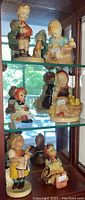 Six figurines displayed on glass shelves showing Hummel and Hummel-style ceramic children figurines in traditional dress with animals and objects.