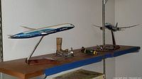 Overall view of the model planes on a wooden shelf, showing 2 Boeing Dreamliner 787 models on metal stands, 3 smaller prop fighter planes, and a small wooden boot figurine.
